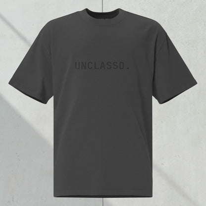 UNCLASSD CORE Oversized Tee in faded black with tonal “UNCLASSD.” chest print, ghost mockup against dark concrete background.