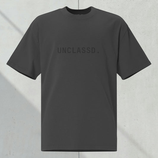 UNCLASSD CORE Oversized Tee in faded black with tonal “UNCLASSD.” chest print, ghost mockup against dark concrete background.